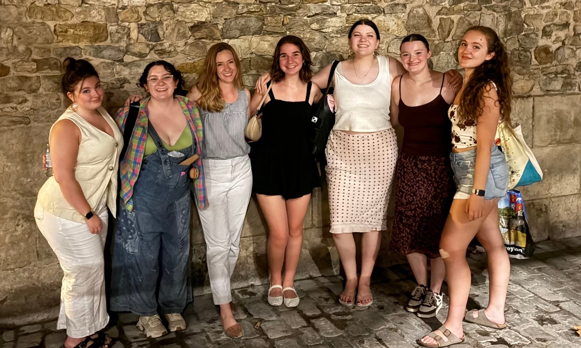 Six women stand closely together in front of a stone wall, posing for a group photo. They are dressed in casual summer clothing and smiling at the camera.