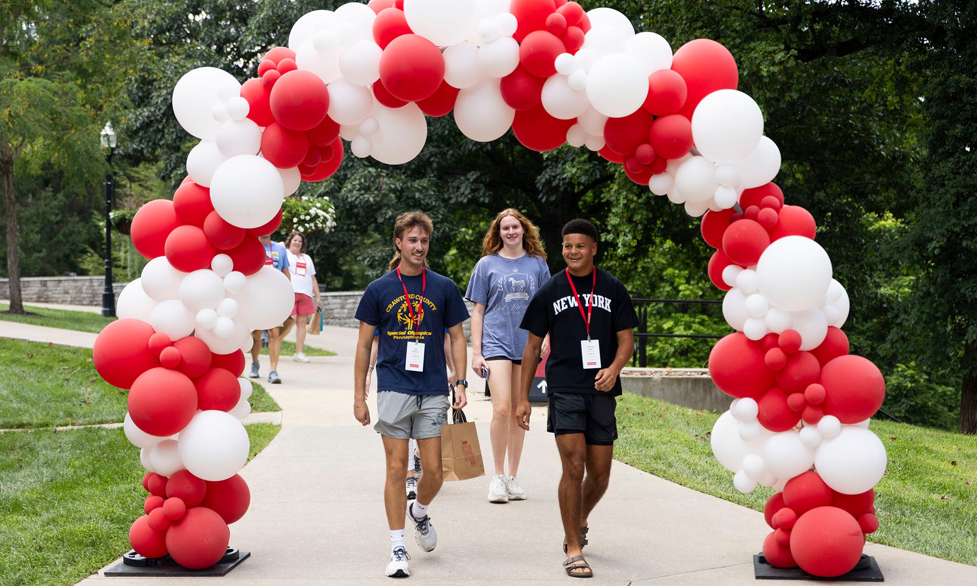 Three people walk under a red and white balloon arch on a sidewalk outdoors, with greenery in the background.