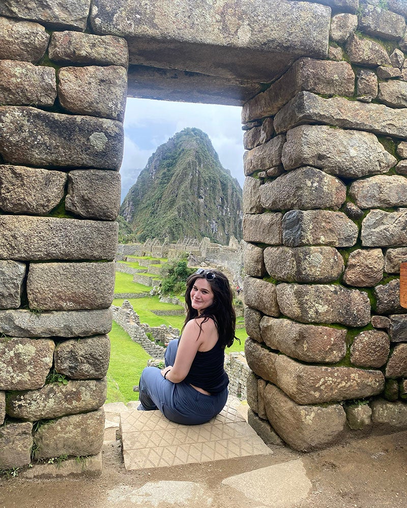 A woman sitting in a stone window structure overlooking a mountain.
