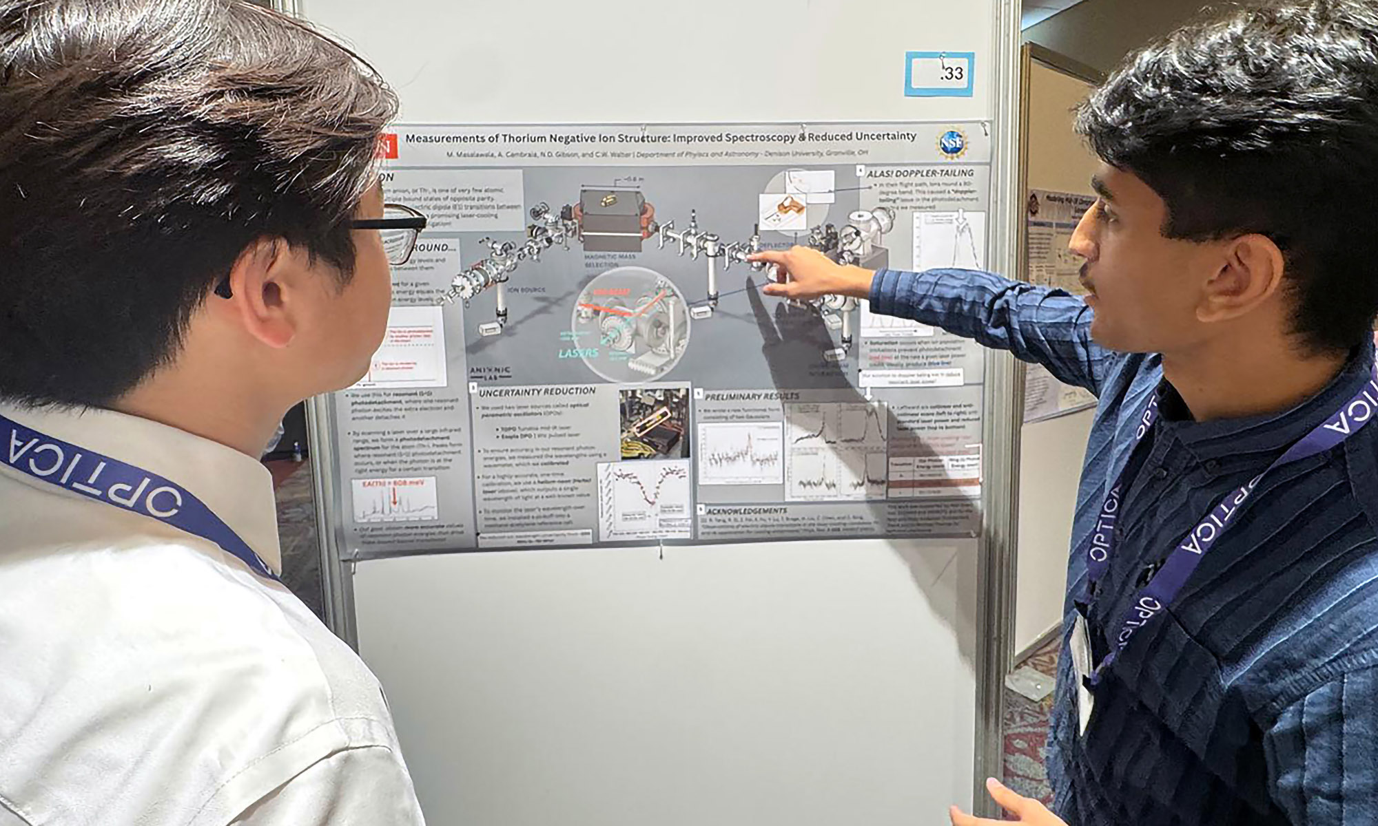 Two students stand in front of a research poster, one pointing to a diagram while explaining it to the other.