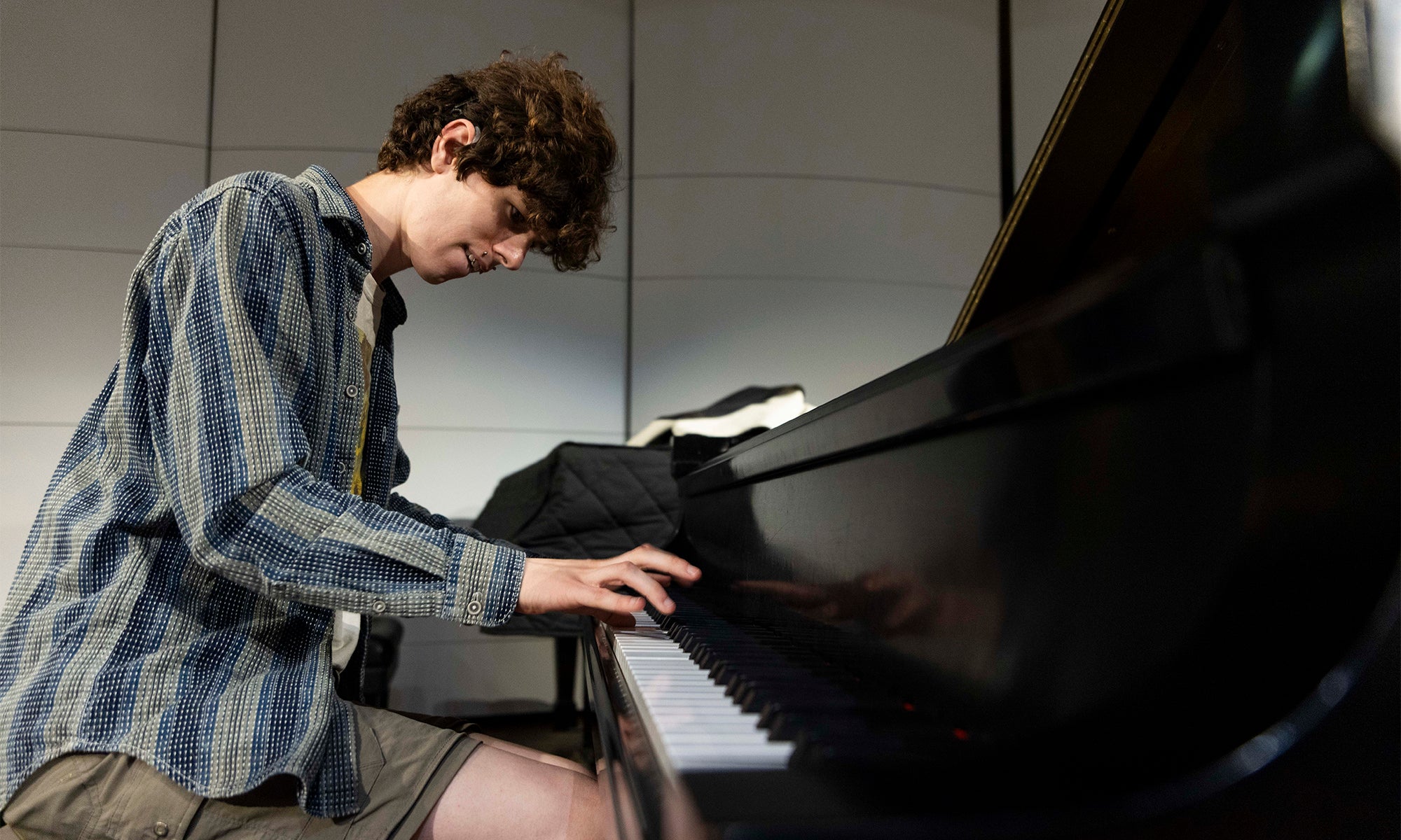 A person with curly hair and a striped shirt sits at a grand piano, playing music. The piano lid is open and sheet music rests on the stand. The background shows light-colored wall panels.