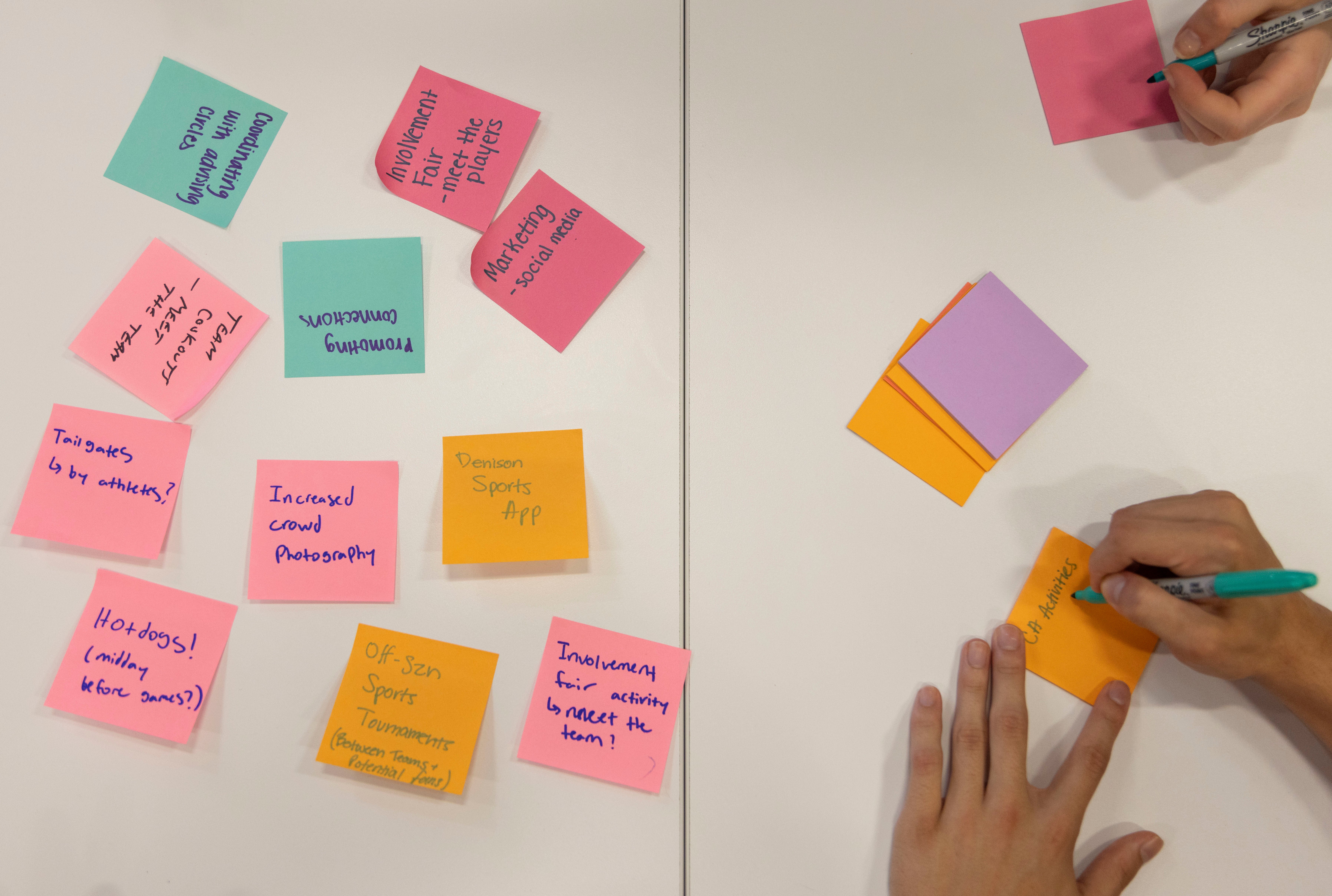 Several colorful sticky notes with handwritten text are spread on the left side of a white table, while on the right, a person writes on a stack of sticky notes with a green pen.