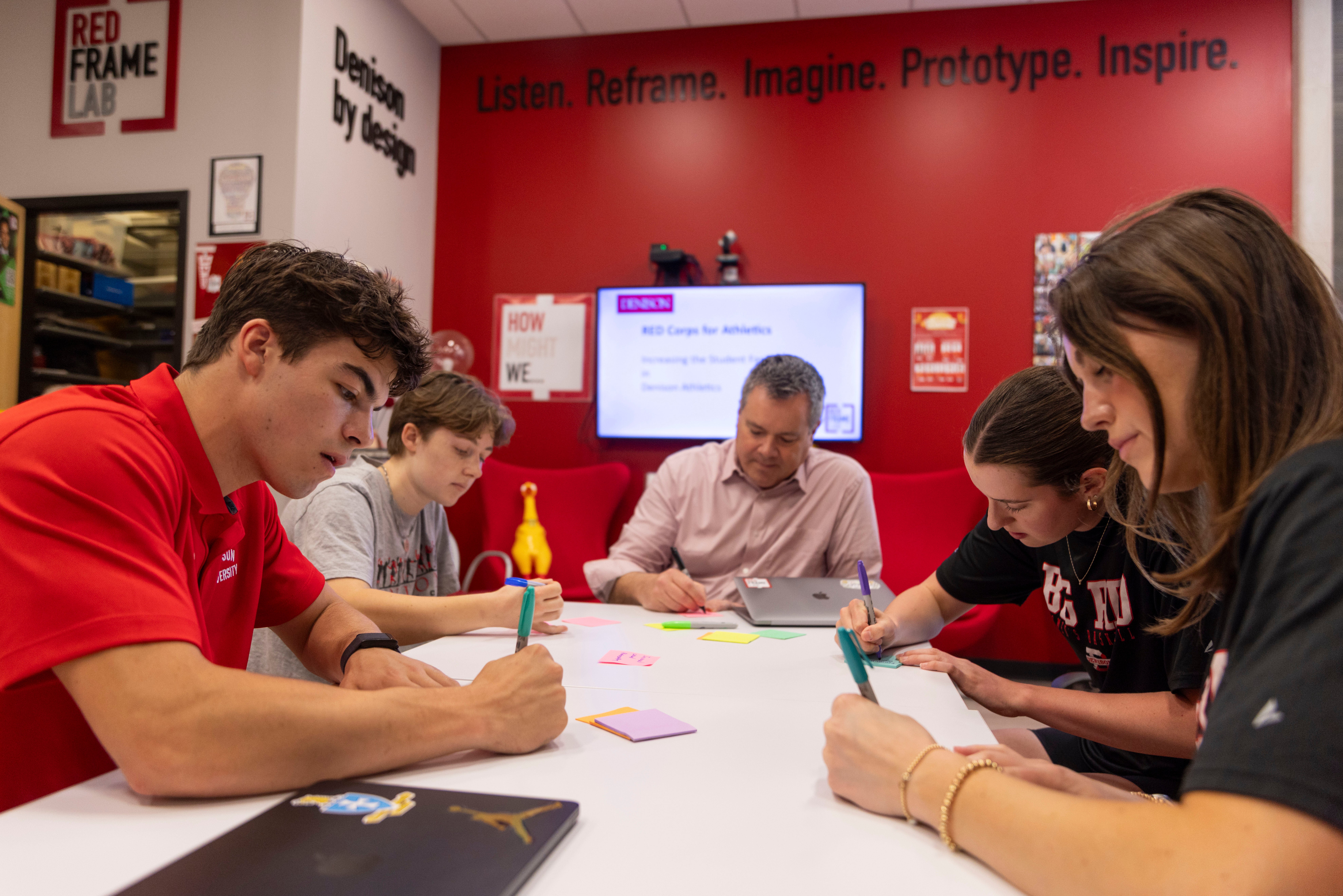 Four students and one adult sit around a white table writing on paper in a classroom with red walls. A screen, posters, and sticky notes are visible in the background. 