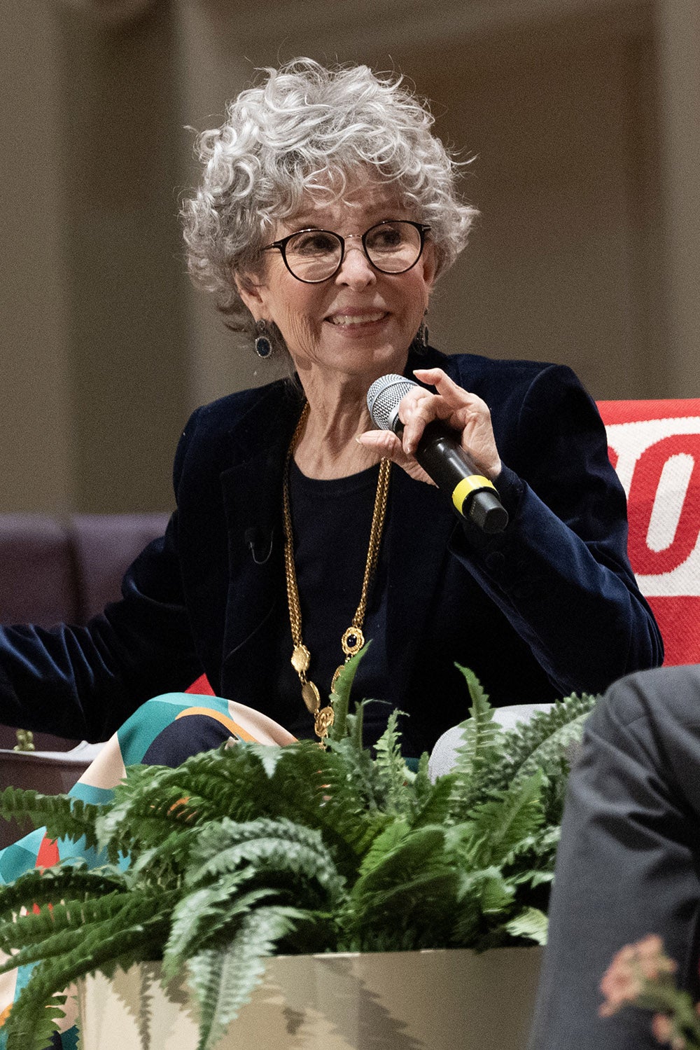 An older woman with curly gray hair and glasses sits on a stage, holding a microphone. She is wearing a dark blazer, a long necklace, and is seated behind a potted plant.