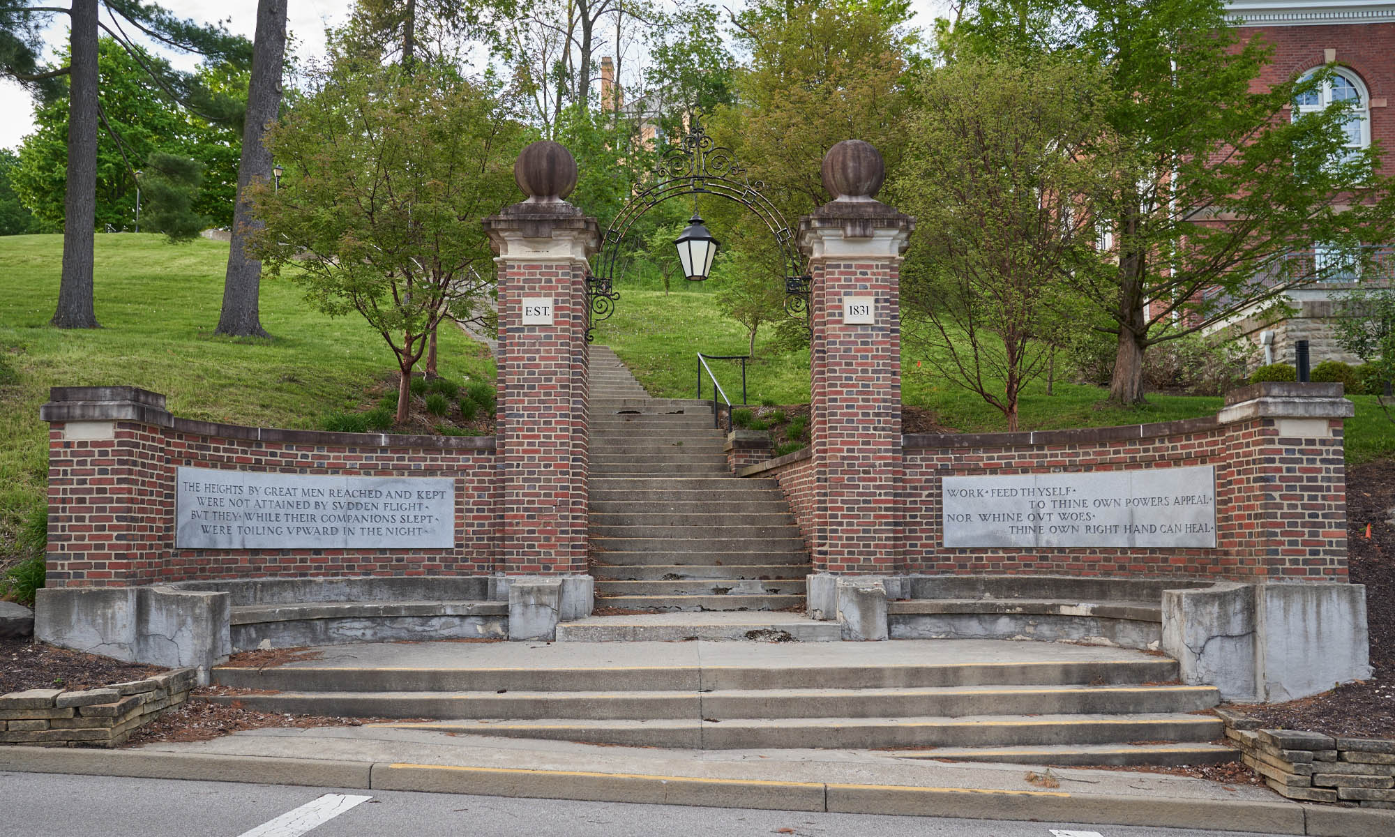 Two brick pillars flanked by brick walls engraved with words; steps lead up from a street to the pillars, which serve as a walking entrance, and continue up a large green hill dotted with trees and buildings near the top.