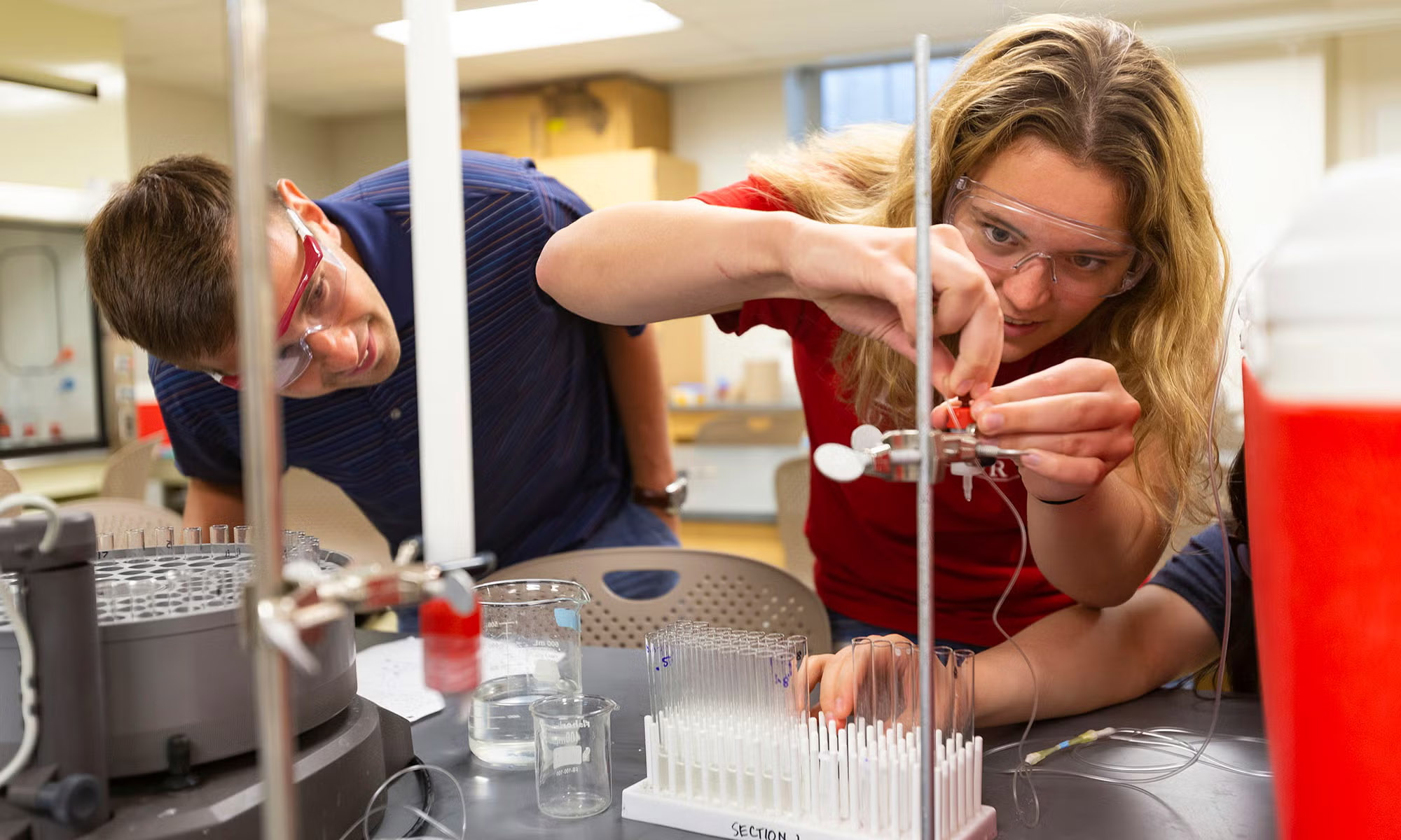 Two people wearing safety goggles conduct a science experiment in a laboratory, using laboratory glassware and equipment on a table.