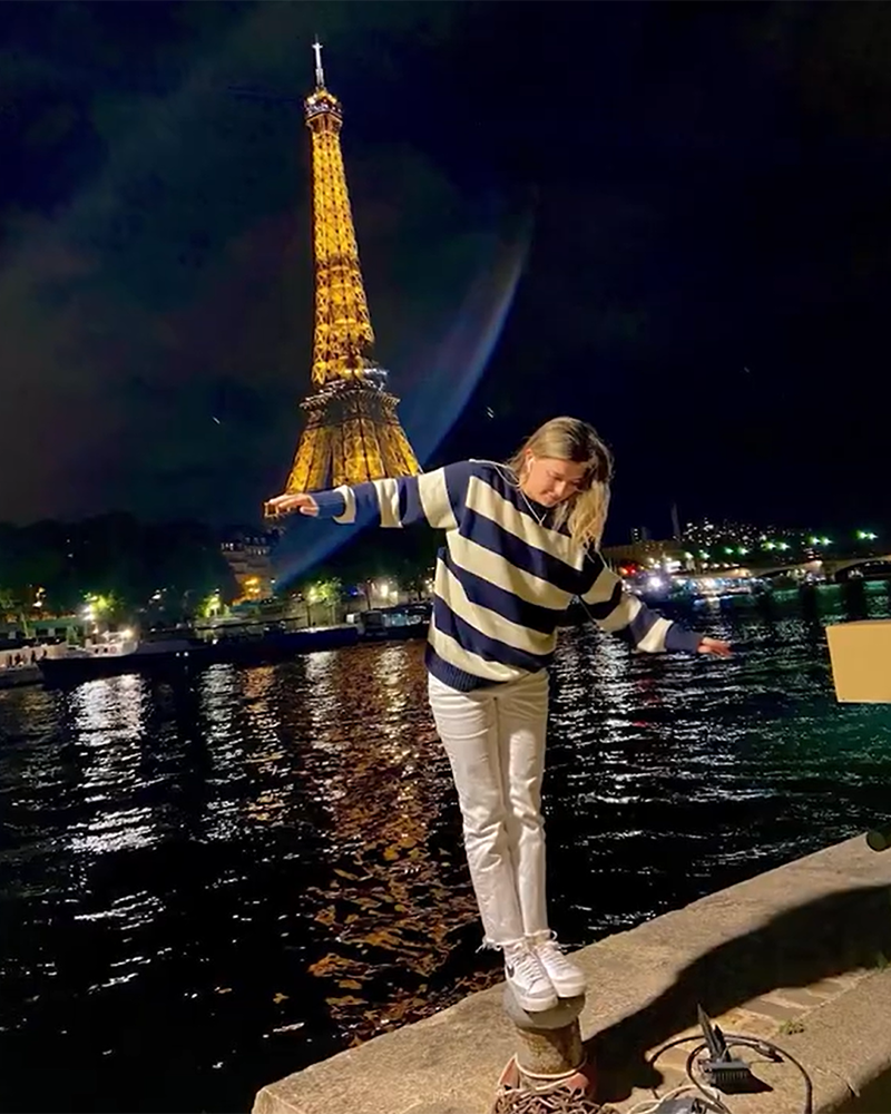 A person balances on a low wall by the Seine River at night with the Eiffel Tower illuminated in the background.