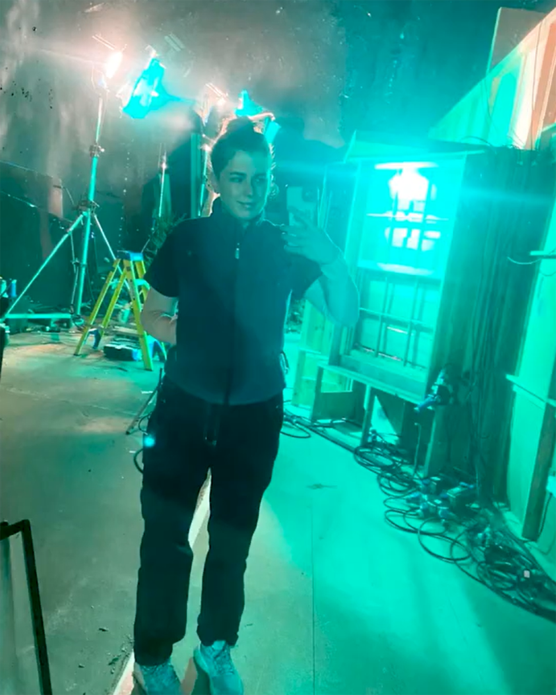 A French and cinema major takes a selfie in a backstage area, surrounded by lighting equipment, cables, and set structures—all illuminated by teal lighting.