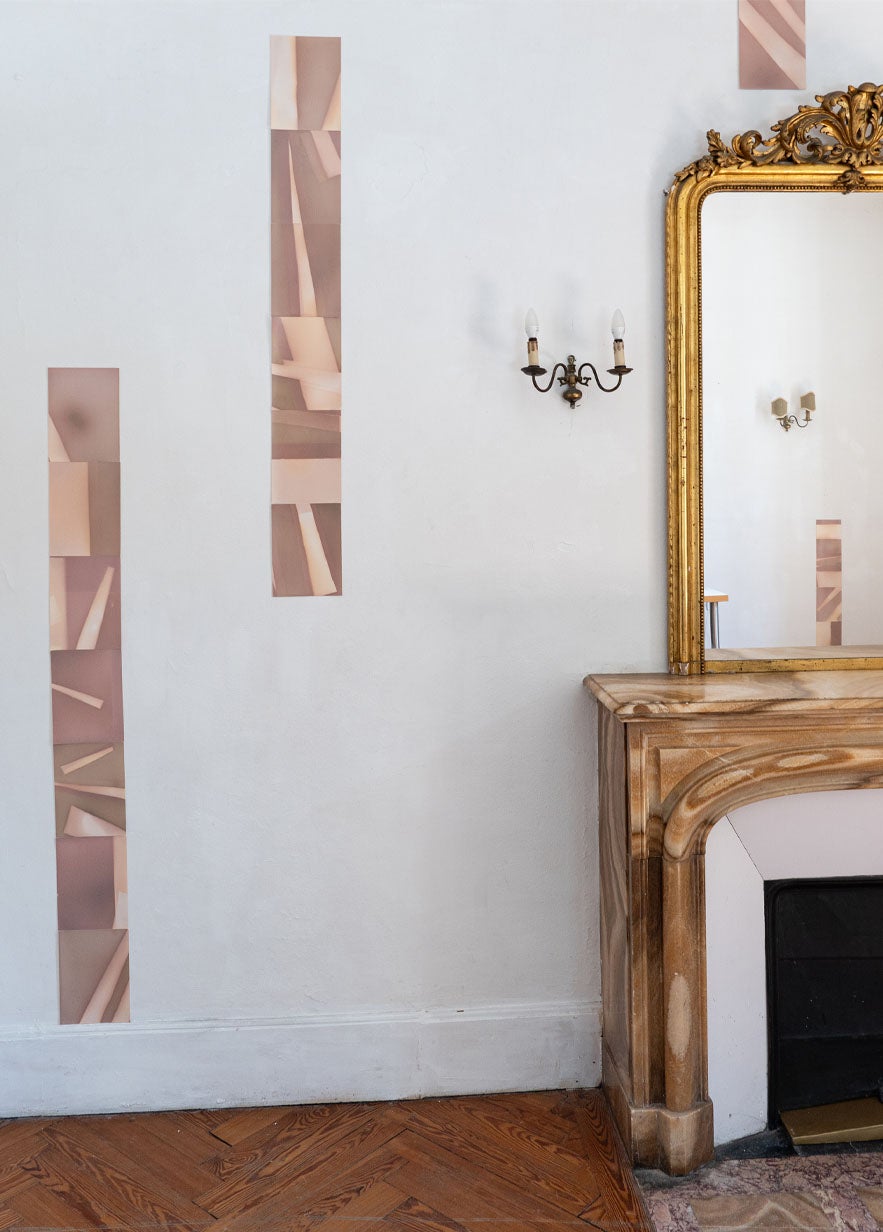 A marble fireplace with a gold-framed mirror above it, wall sconce, and three vertical abstract wall art panels on a white wall.