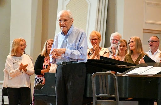 William Osborne stands and speaks in front of a piano, with people standing behind him applauding