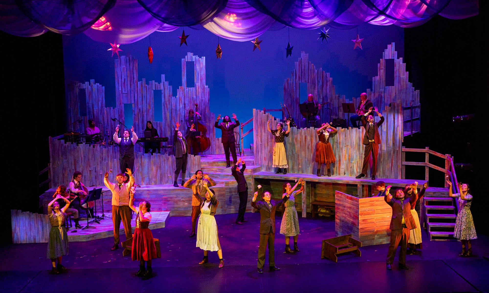 Stage scene of a musical theater performance with actors in period costumes, some on platforms and others on stage, set against a colorful cityscape backdrop under dramatic lighting.