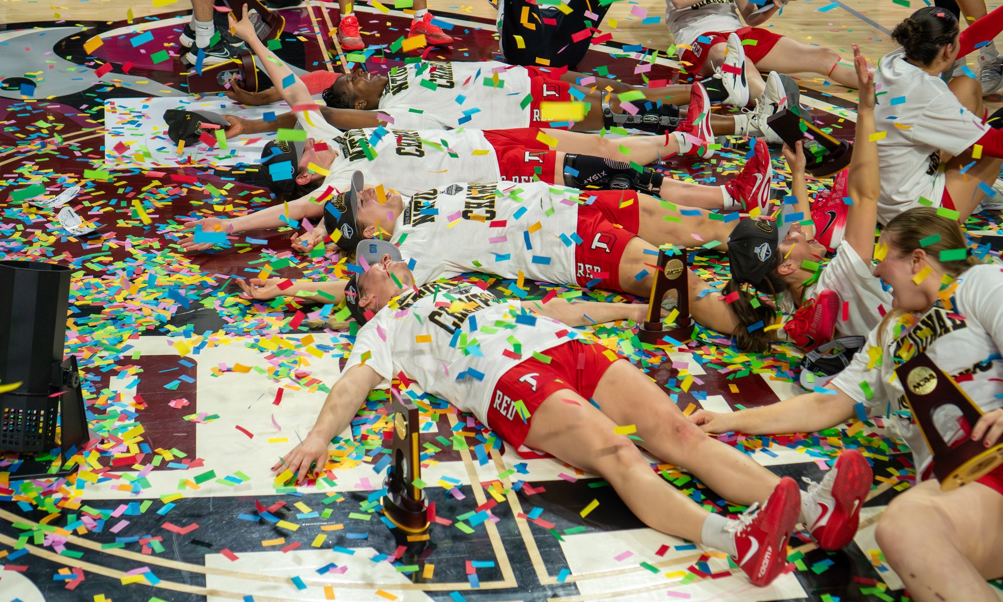 Women's basketball players making "snow angels" in a sea of confetti on a basketball court floor.