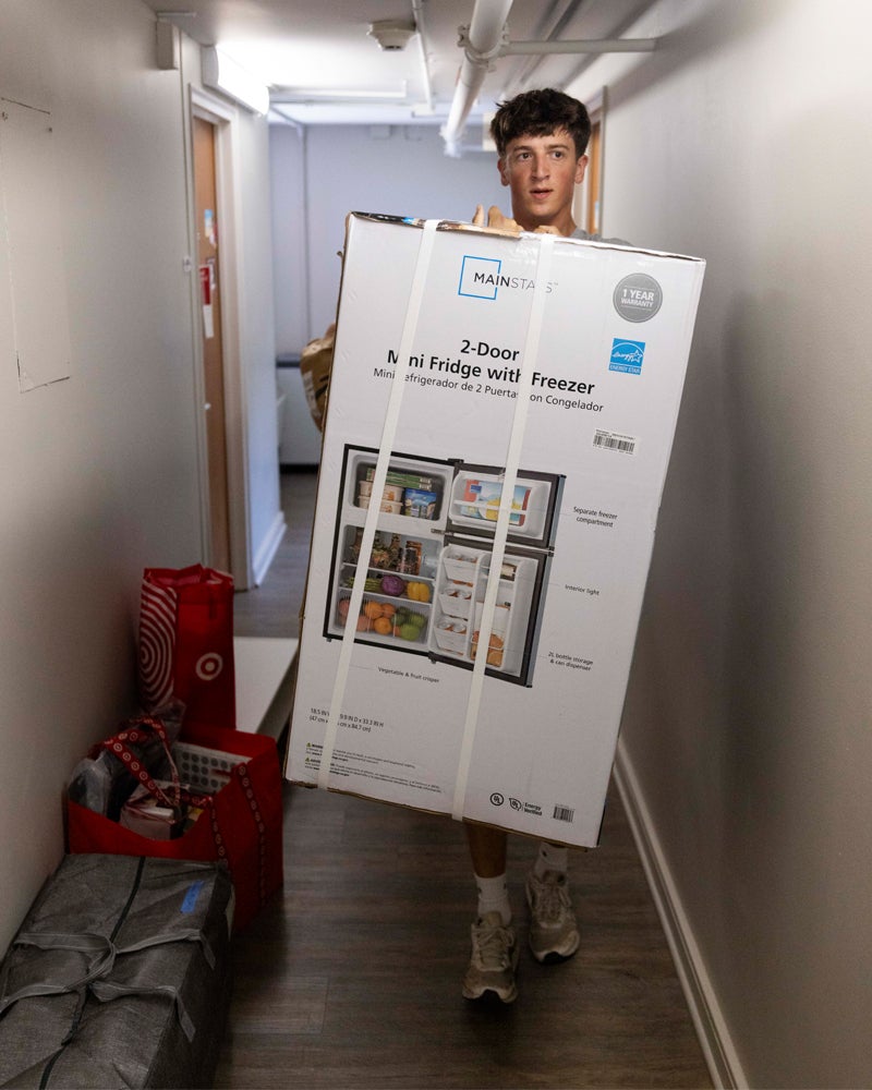 A person stands in a hallway carrying a large boxed 6-shelf wire closet organizer. Red bags and silver storage bins are on the floor nearby.