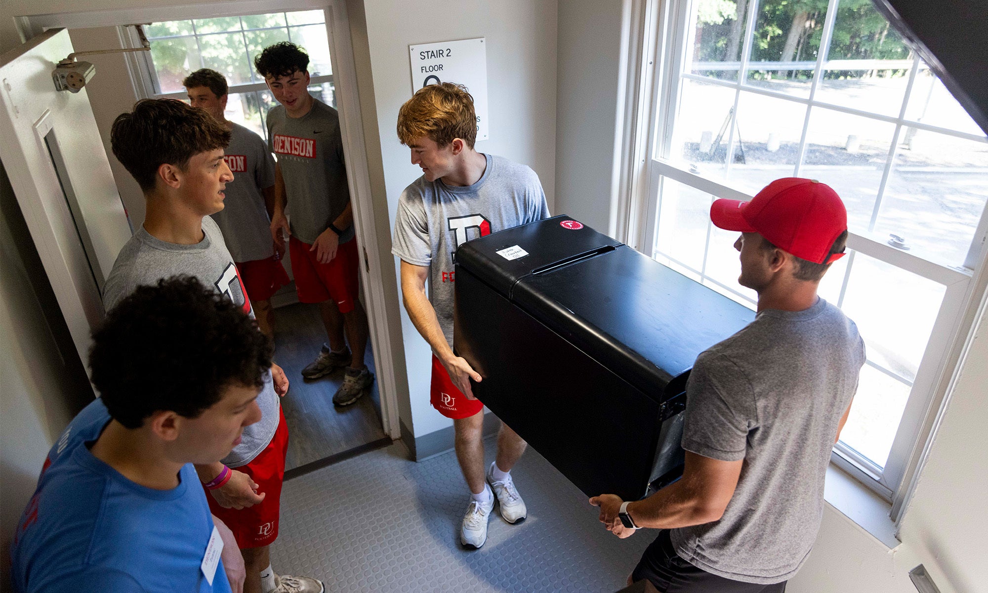 Two young men carry and guide a mini fridge through a doorway in a brightly lit room, with sunlight coming through a window as others help guide them.