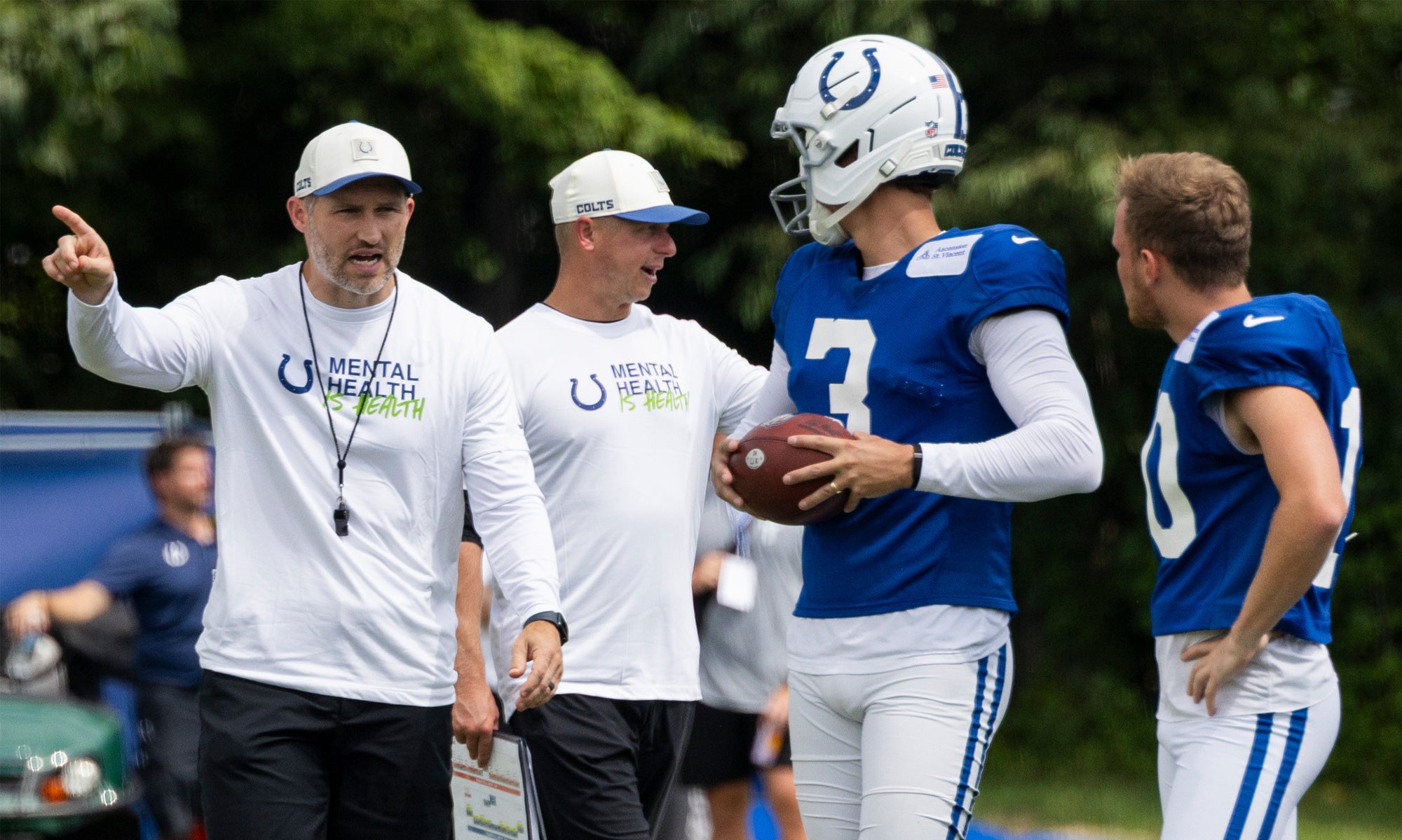 Football coaches give instructions to players in blue Indianapolis Colts uniforms during a practice session outdoors.