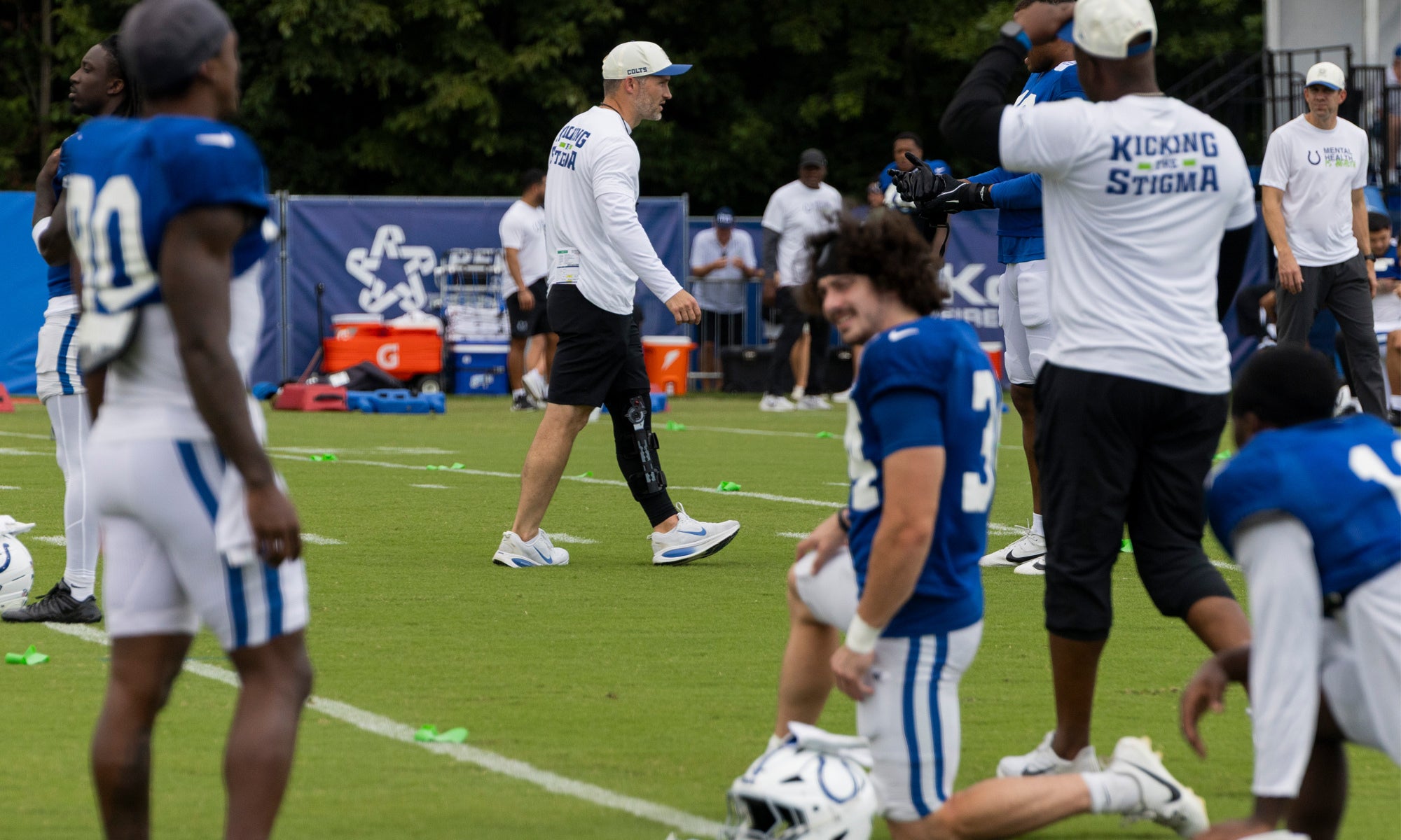 Football players in blue and white uniforms stretch on a grassy field during practice, with coaches and staff standing nearby.