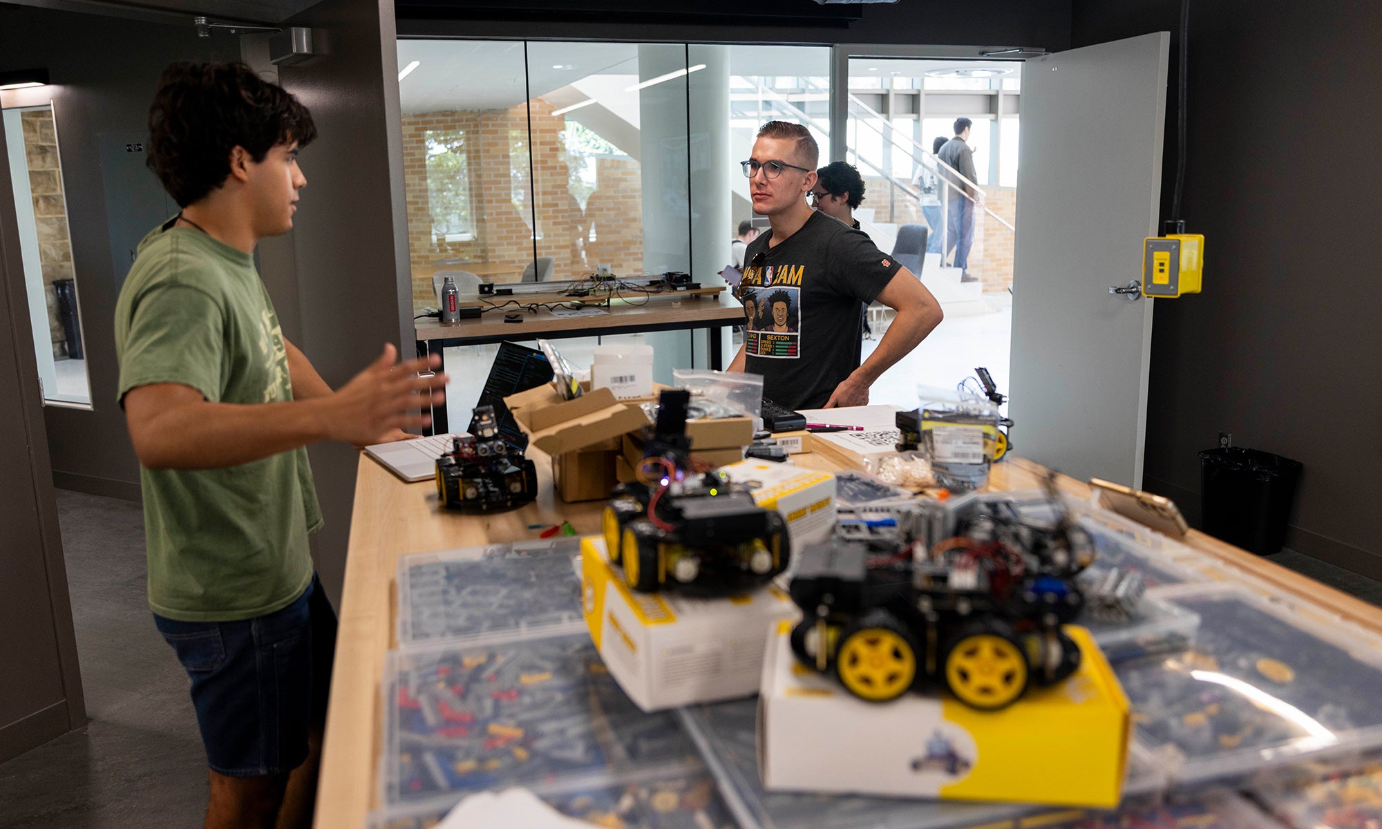 Two people discuss at a workspace table covered with robotics kits and components, with additional people and equipment visible in the background.