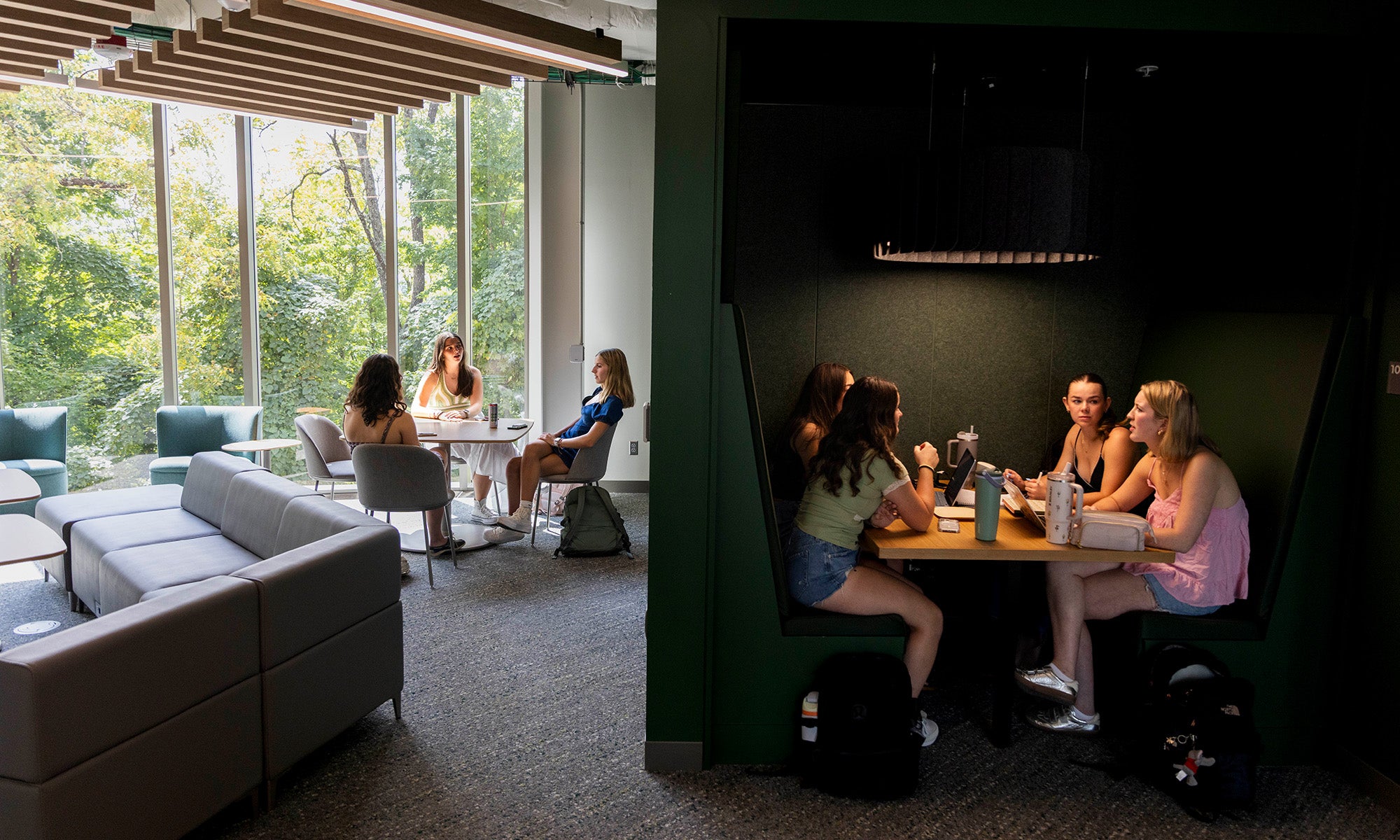Students sit and talk in groups, some at a round table near large windows, others in a booth, inside a modern, well-lit lounge with greenery visible outside.