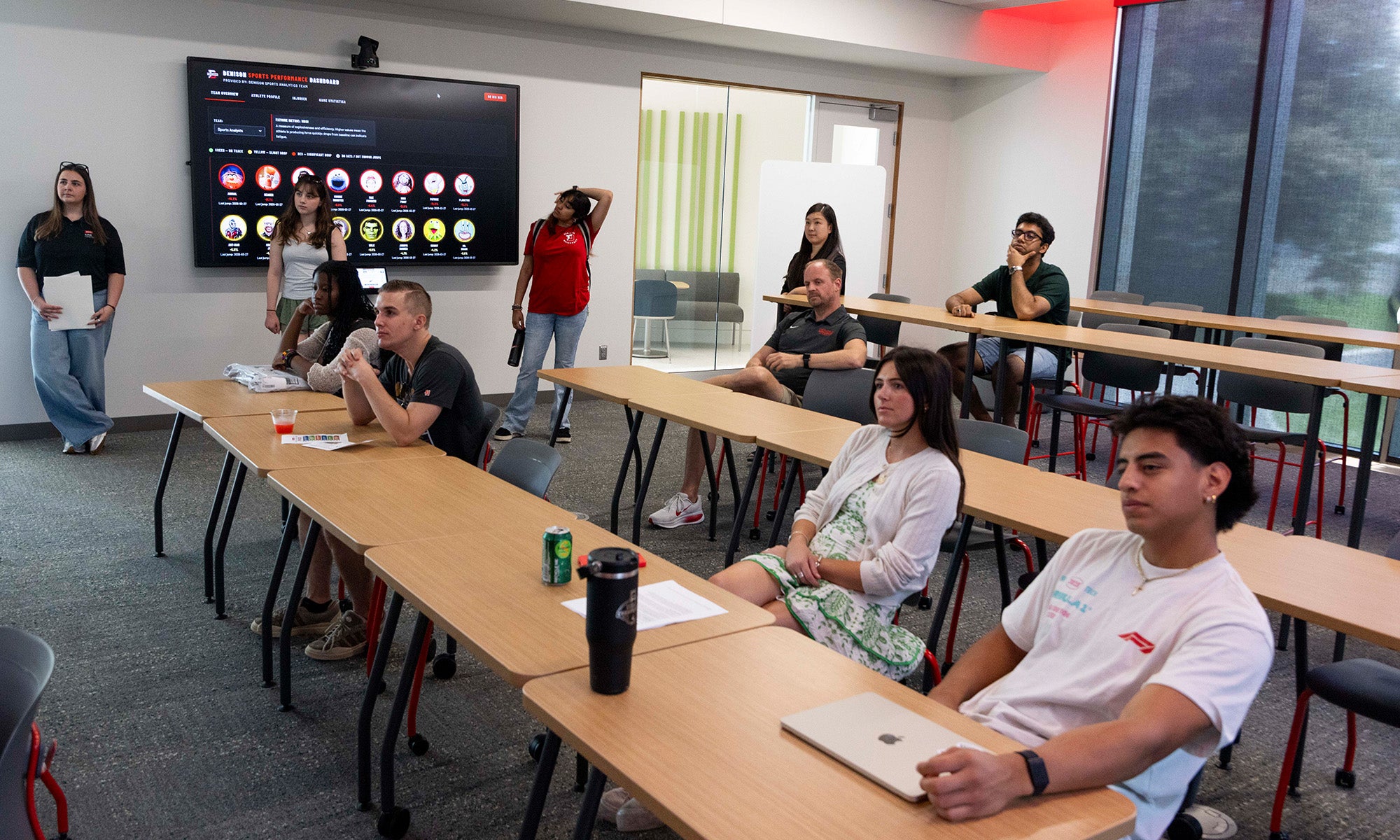 Students sit at desks in a classroom, some with laptops, while a large screen displays icons at the front and an instructor stands nearby.