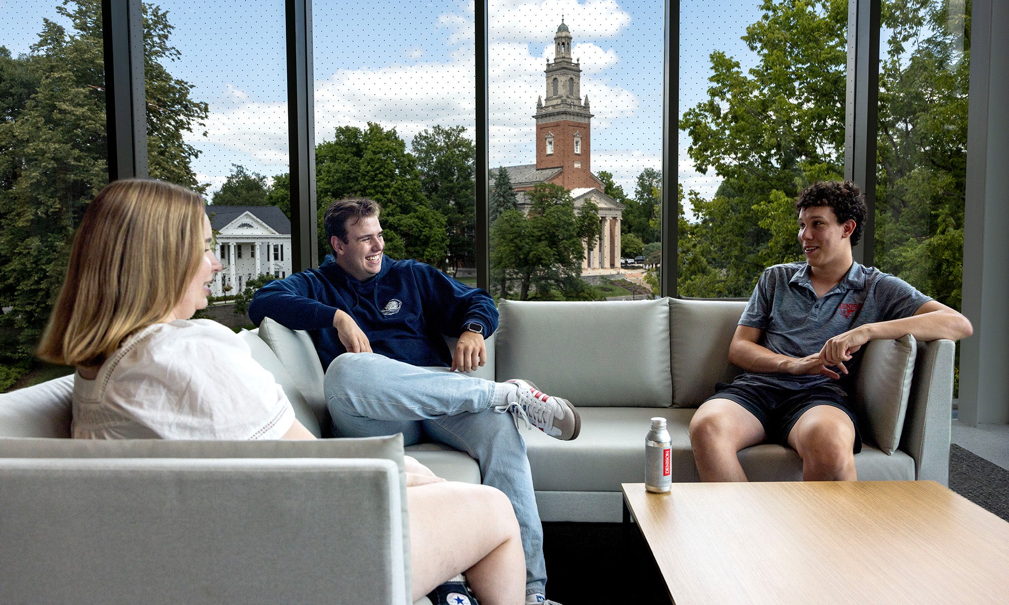 Three people sit on couches in a lounge with large windows, talking. A building with a tall clock tower is visible outside among trees.