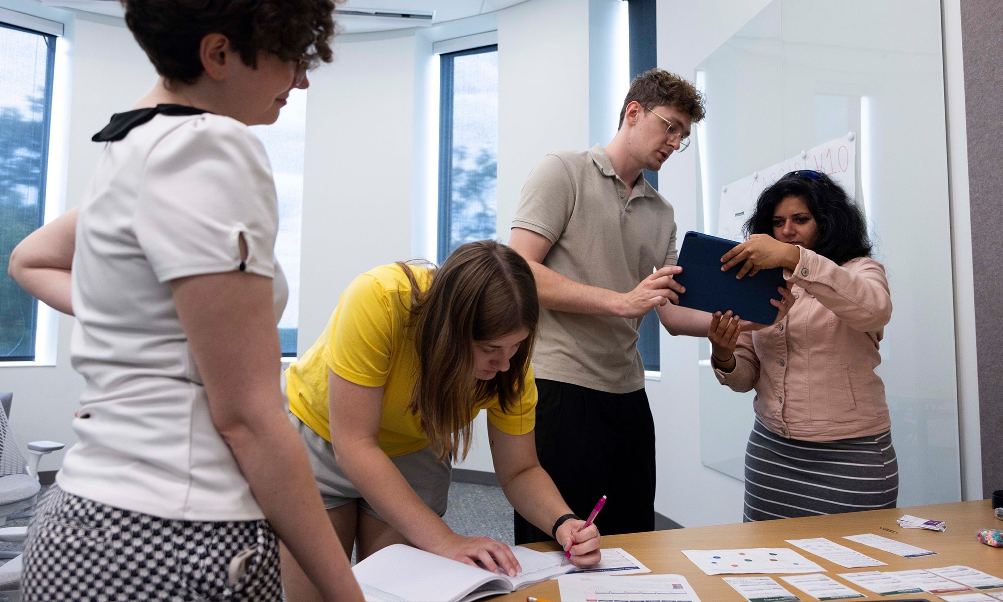 Four people work together at a table; one writes in a notebook, two examine a tablet, and one stands nearby. Papers and cards are spread out on the table.