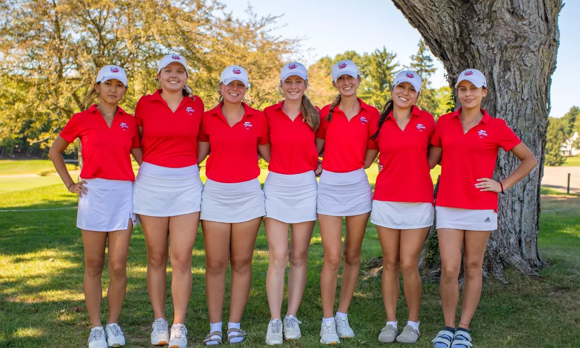Seven young women in matching red shirts, white skirts, and white caps stand side by side outdoors on grass near a large tree, posing for a group photo.