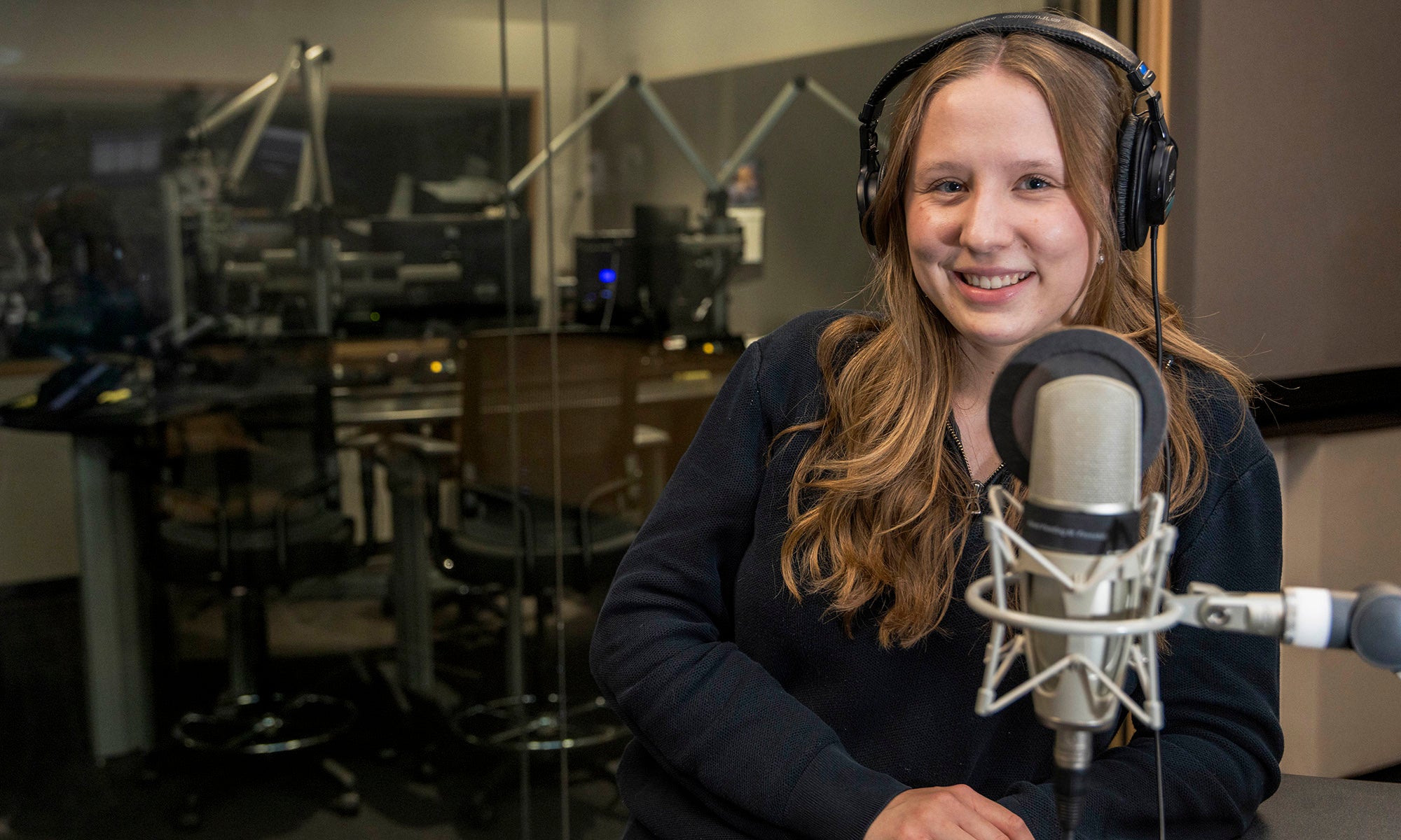 A woman with long hair wearing headphones sits in front of a microphone in a radio studio.