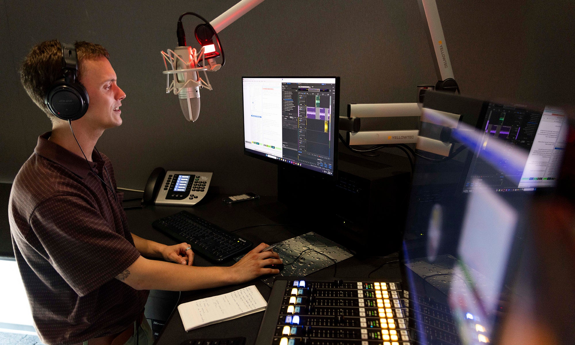 A person wearing headphones speaks into a microphone in a recording studio with multiple monitors, a soundboard, and other audio equipment.
