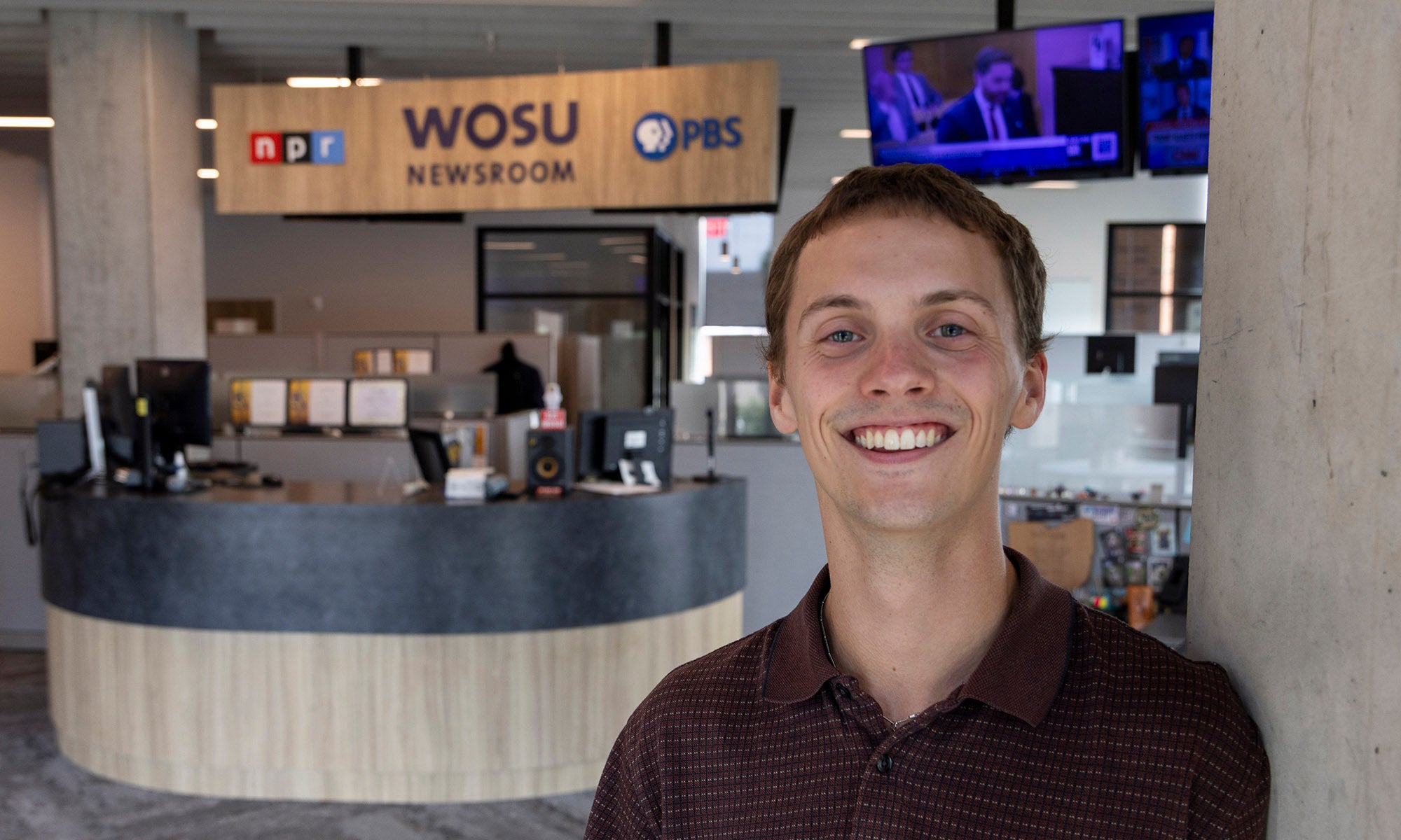 A young man smiles in front of a WOSU newsroom reception area with a PBS and NPR sign and TV screens in the background.