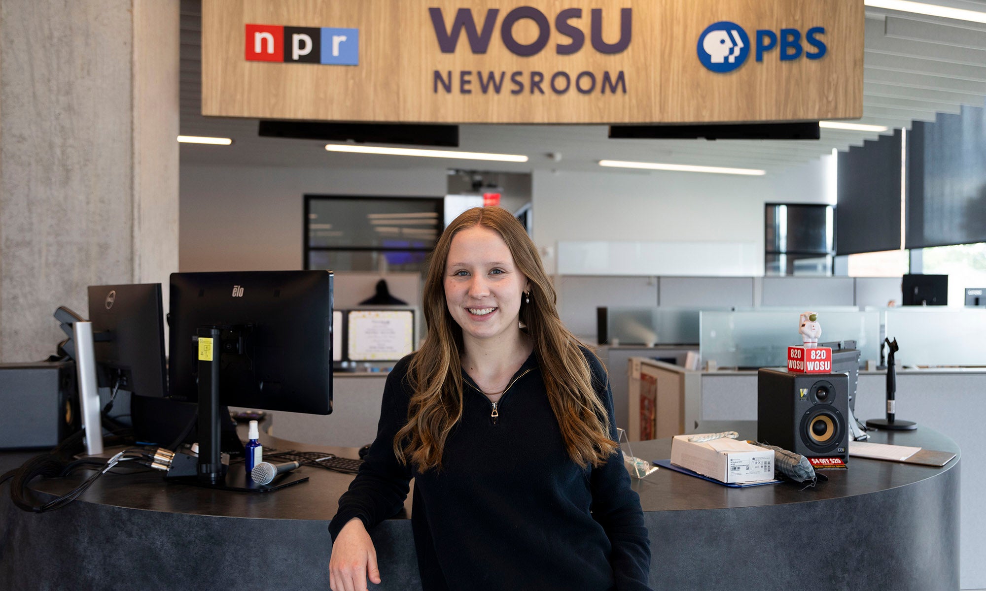 A woman stands in the WOSU newsroom, with NPR and PBS logos displayed above the desk and various office equipment in the background.