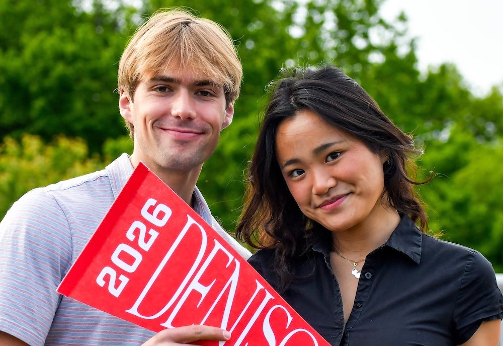 Two people stand outdoors holding a red pennant that says Denison 2026. Both are smiling and posing for the photo, with green trees in the background.