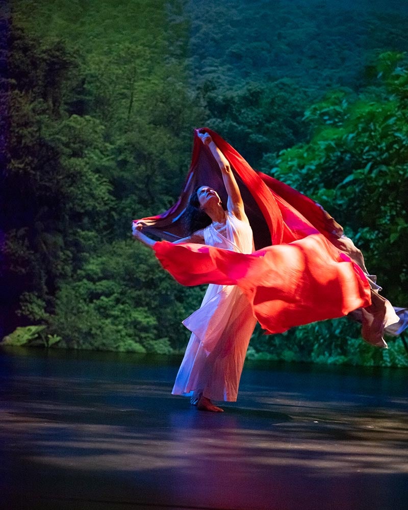 A dancer in a flowing white dress moves gracefully, holding and swirling a large red fabric against a backdrop of green foliage.