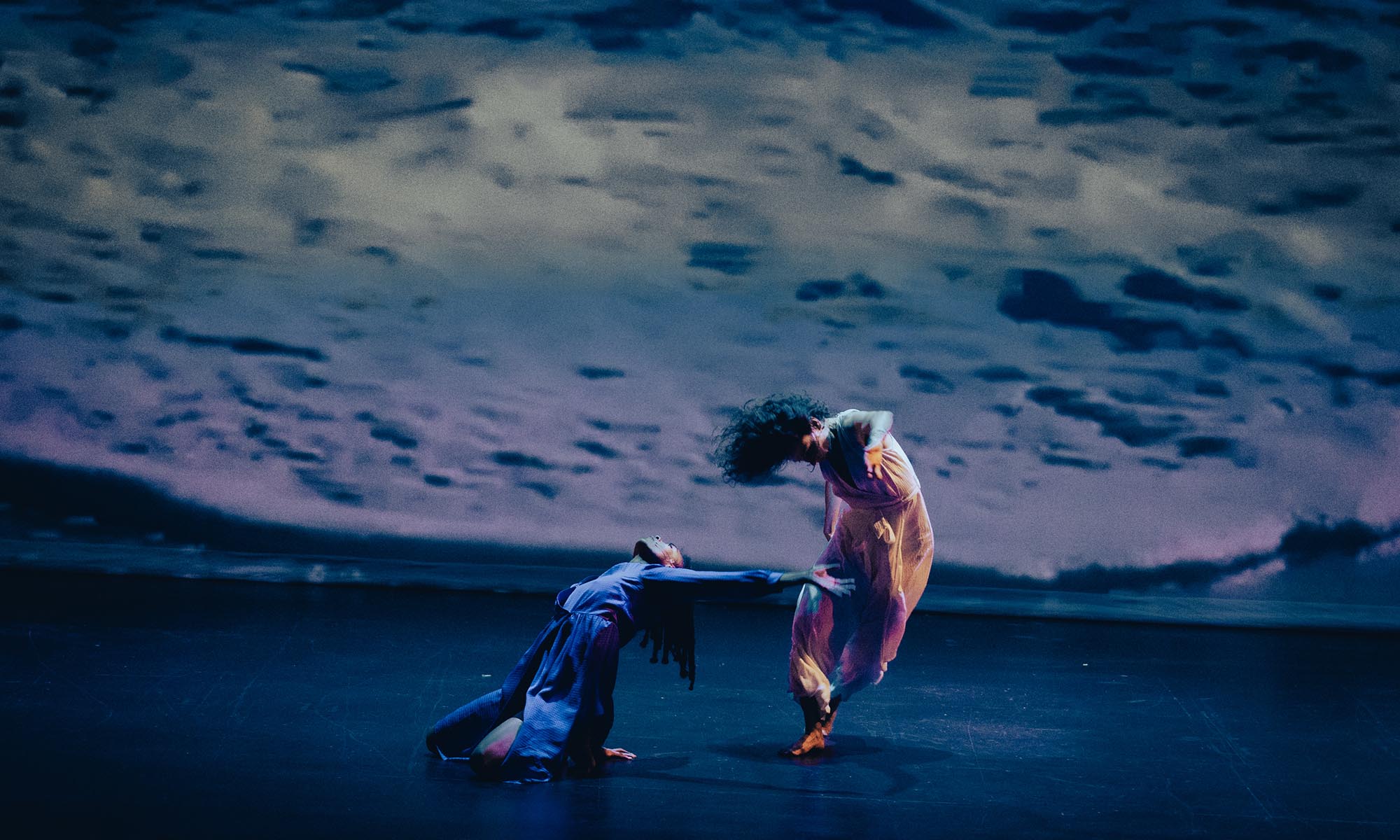 Two dancers moving together on a blue-lit stage against the backdrop of an ocean tide.