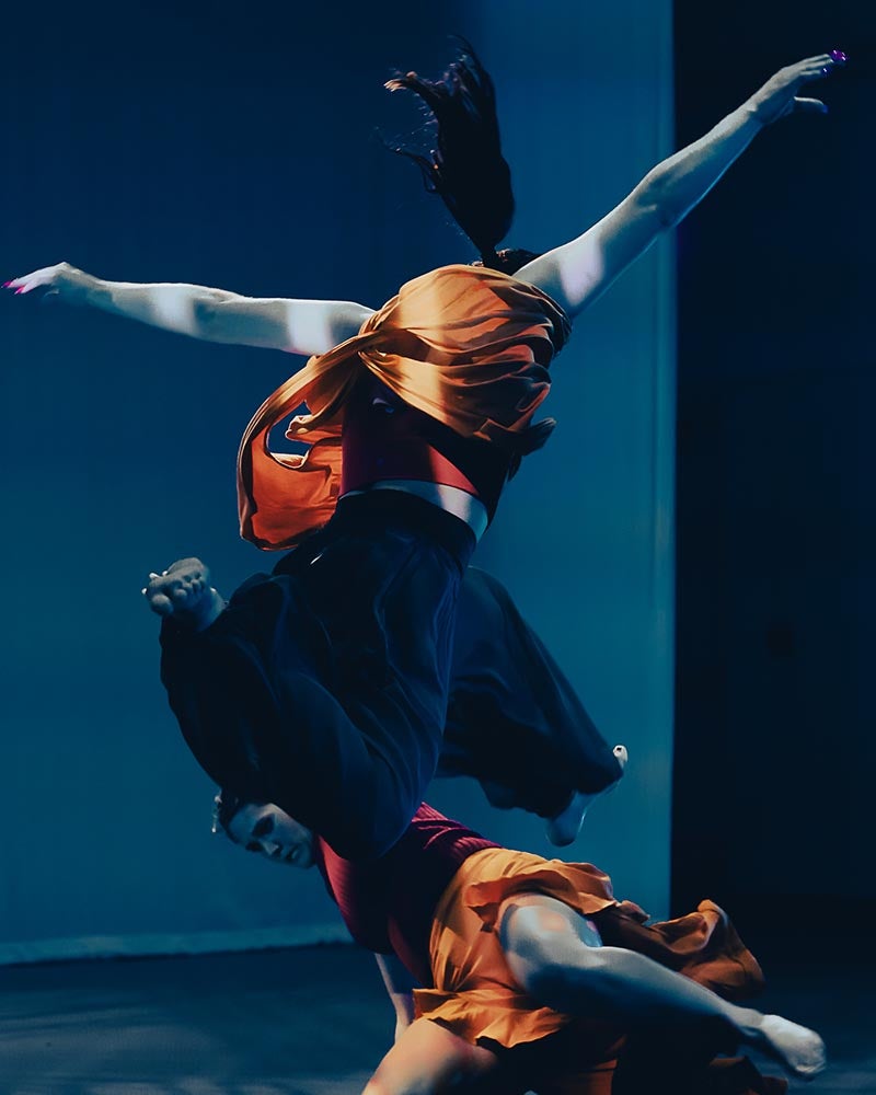 A dancer in dark blues and orange flowing clothes dances on a blueish-tinted stage.