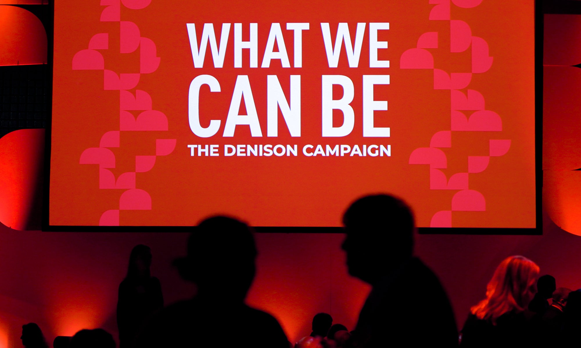 People mingle in front of a sign that says "What We Can Be."