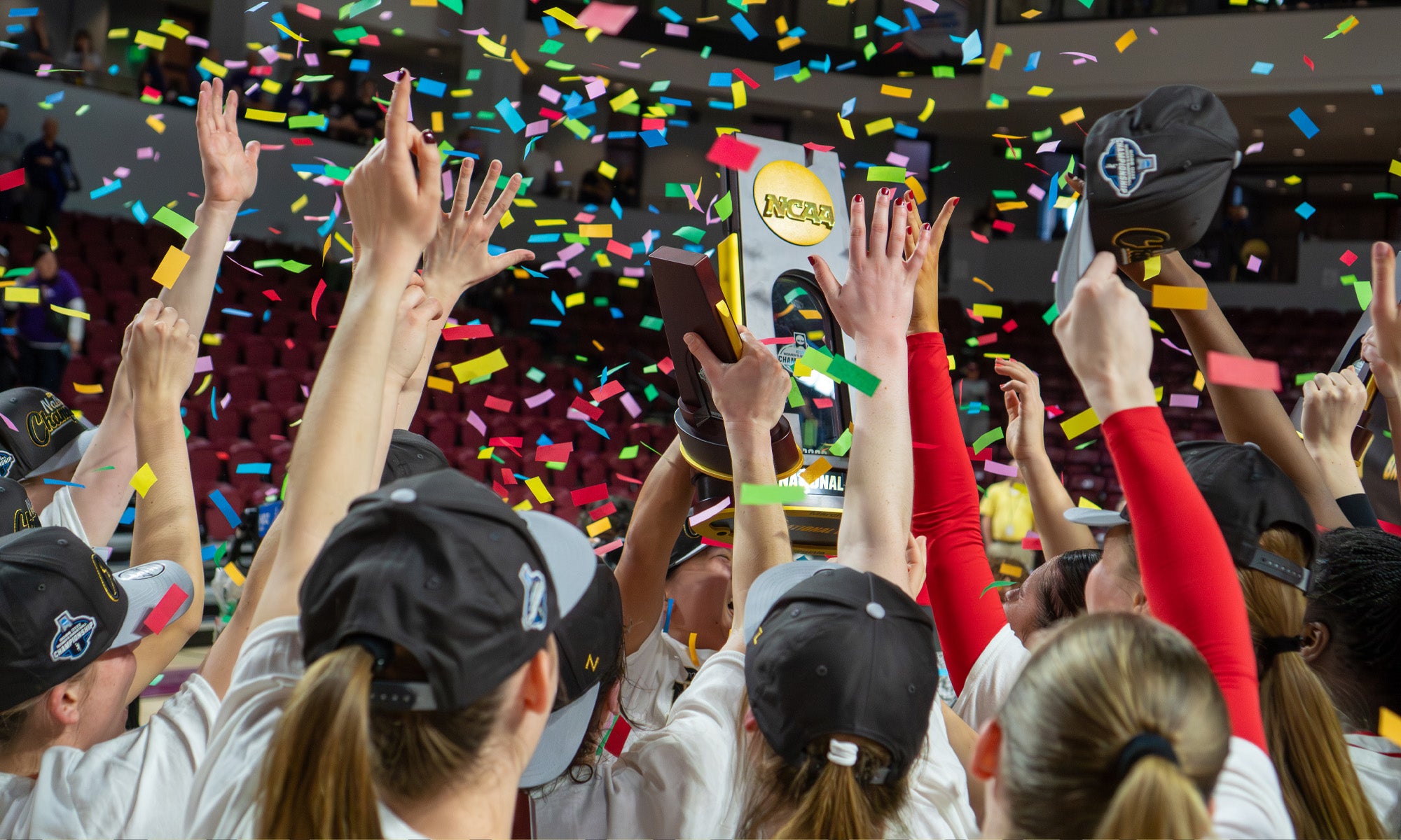 A sports team wearing hats raises a trophy together amid falling confetti in an indoor arena, celebrating a championship victory.