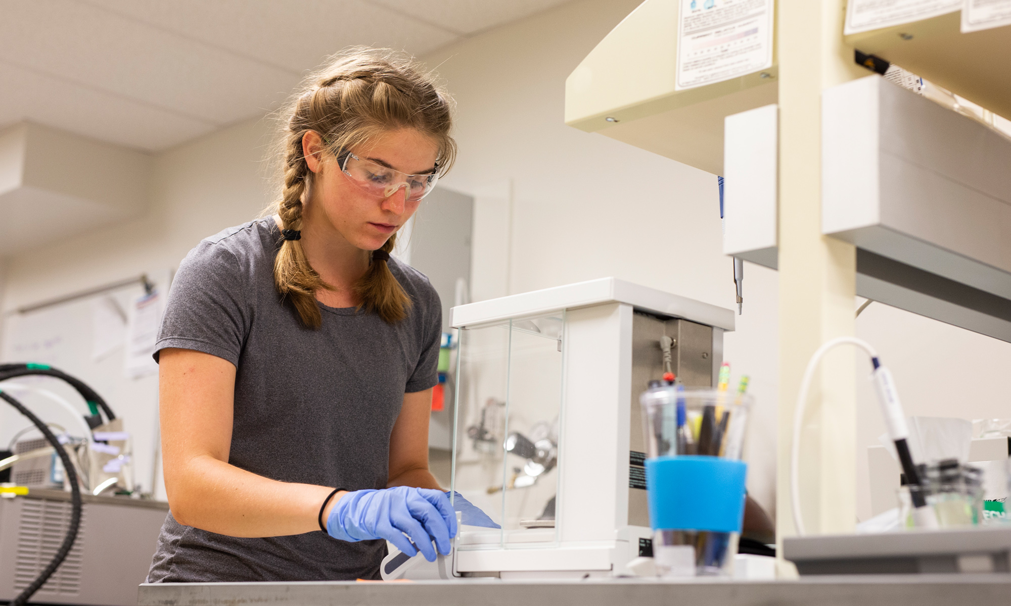 A biochemistry student wearing goggles and gloves performs an experiment in a classroom lab.
