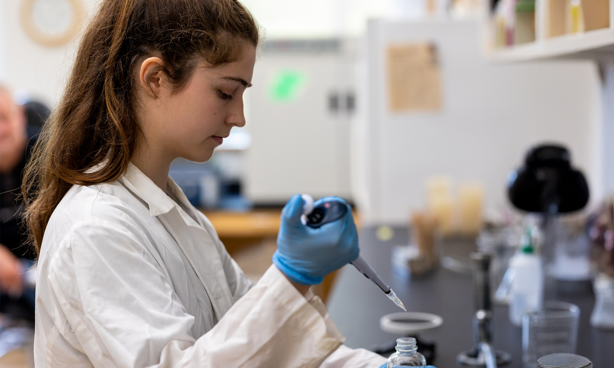 A biology student in a lab; she wears a lab coat and gloves and holds a pipet.