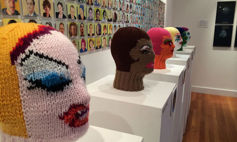 Knitted faces with portraits hanging on the wall behind them