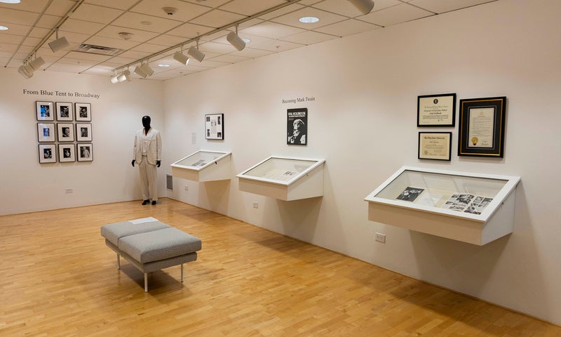 A museum exhibit room at the Denison Museum with framed photographs and documents on white walls, angled display cases beneath, a gray bench in the center, and a mannequin dressed in a white coat near the back.