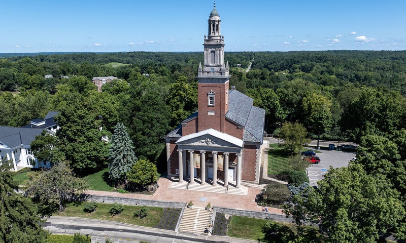 A red brick church with a tall steeple, white columns, and a clock tower, surrounded by trees and a parking lot on a clear, sunny day.