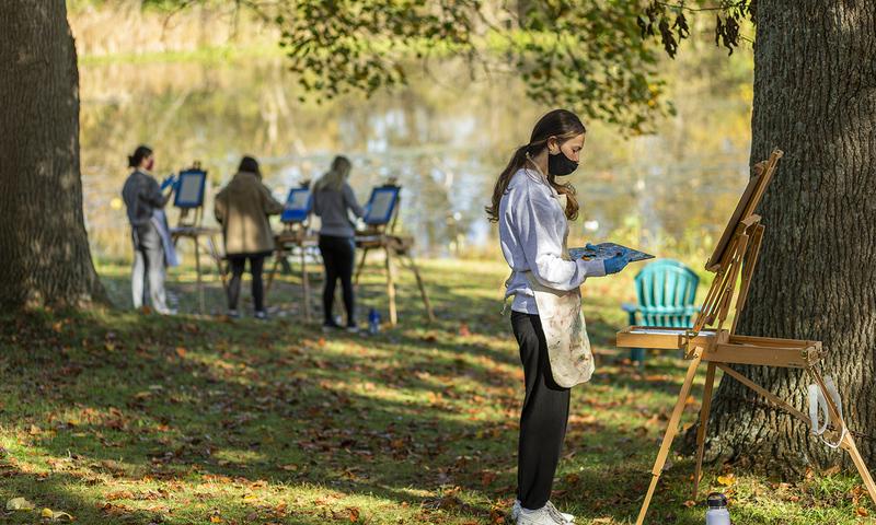 Students paint on easels outdoors while wearing masks