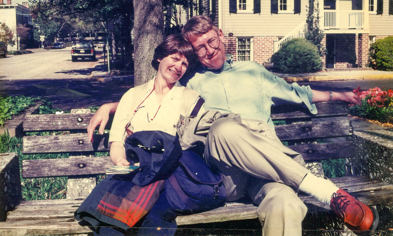 A man and a woman sitting on a park bench on a warm day. His arm is around her and she is leaning into him.