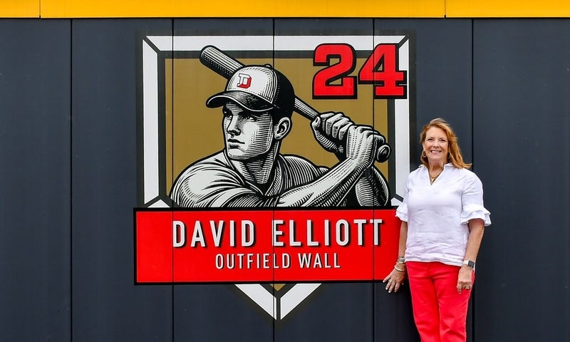 A photo of Lisa Elliott standing by the newly unveiled David Elliott outfield wall graphic.