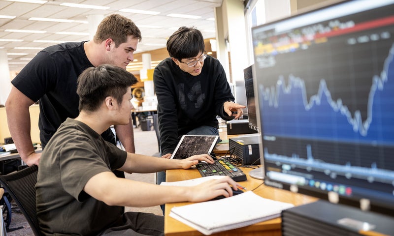 Three people in a computer lab look at a monitor together as one person points to an unseen data point; another monitor displaying stock market graph sits slightly out-of-focus in the foreground..