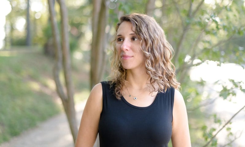 A woman with wavy light brown hair wearing a sleeveless black top stands outdoors, looking slightly to the right, with trees and greenery in the background.