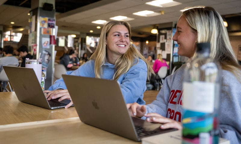 Two women sit at a table with laptops, smiling and talking to each other in a busy indoor setting.