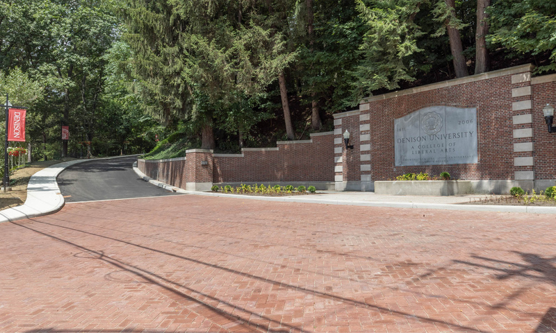 Photo of Presidents' Drive from the main entrance; brick pavers, framed on the right by an ornate brick and stone gateway with a Denison University sign lead to a newly paved, tree-lined roadway and sidewalk that winds up a wooded hill, dotted with sign posts featuring Denison banners.