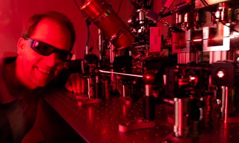 A man in dark protective glasses smiles next to a scientific piece of equipment emitting a laser in a red-hued room.