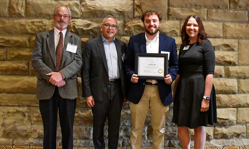 Four people stand in front of a stone wall; the second person from the right holds a framed certificate while the others stand beside him, smiling.